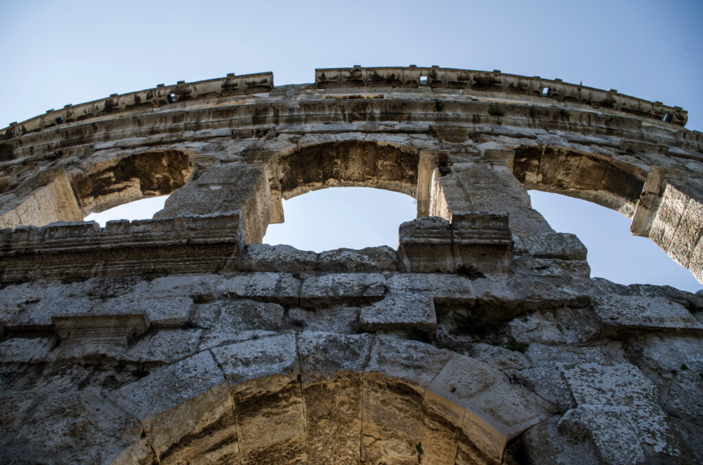 Upward view through ancient Roman amphitheater arches showing keystone architectural principle where multiple tiers of weathered stone arches stack to bear weight across centuries